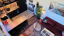 A modern, open-concept living and dining area with a red sofa, coffee table, round stool, and kitchen counter. The space features wooden flooring, patterned rugs, and a potted plant near the window.