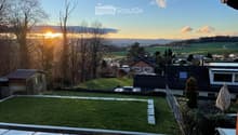 View from a balcony or terrace showing a grassy yard, surrounding houses, trees, and a scenic sunset view of hills and fields.