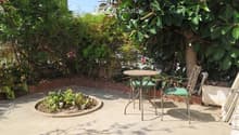 A small outdoor patio with a round table and two chairs, surrounded by lush greenery and plants.