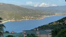 A scenic coastal view with a house on a hillside overlooking the sea, surrounded by lush greenery and distant mountains.