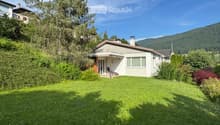 Single-story house with a sloped roof, surrounded by a well-maintained lawn and trees, with a scenic mountain view in the background.