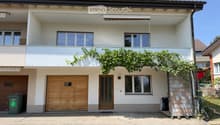Two-story house with white exterior, wooden garage door, and balcony with green vines. Features multiple windows and a covered entrance area.