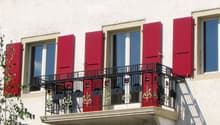 Balcony with black metal railing and decorative elements, red shutters, and large windows on a white building facade.