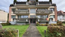 A three-story residential building with balconies, large windows, and a well-maintained garden in front.