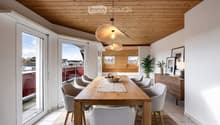 Bright dining area with a wooden table, six chairs, a bowl of fruit, a cabinet with a lamp, and a view of the city through large windows.