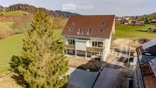 A modern two-story house with a brown tiled roof, large windows, and balconies, situated in a rural landscape with green fields and trees.