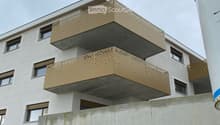 Modern multi-story building with white facade, large windows, and distinctive gold-colored balconies. Features a raised ground floor and a visible garage entrance.