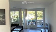 Modern living room with large windows and a balcony, featuring a sofa, coffee table, TV stand, and chairs.