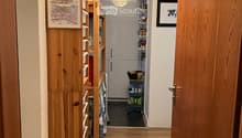 A narrow hallway with wooden flooring, white walls, and a ceiling light. A wooden shelf with a framed picture and storage boxes is on the left, and a partially open door leads to a room with a refrigerator and shelves.
