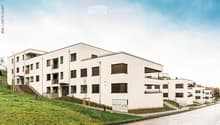 Modern multi-story residential building with white facade, multiple windows, balconies, and surrounding greenery.