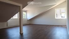 Empty attic room with wooden flooring, white walls, and large windows allowing natural light. The room features a slanted ceiling and a central support column.