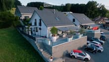 Aerial view of a modern two-story house with a sloped roof, surrounded by greenery and neighboring houses. Features include a raised ground floor, a balcony, and a parking area with several cars.