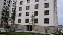 Modern multi-story residential building with white facade, multiple windows, and balconies. The ground level features a landscaped area with grass, small trees, and a paved walkway.