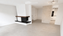 Open-plan living space with a fireplace, white walls, grey tiled floor, and recessed lighting. The room is unfurnished, showing only the fireplace and a doorway leading to another area.