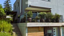 Modern two-story house with grey facade, large windows, balcony, and steps leading to a wooden garage area with bicycles parked nearby.