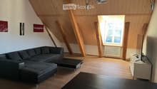 Modern living room with wooden slanted ceiling, large window, grey sectional sofa, coffee table, TV stand, and additional table.