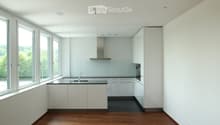 Modern, minimalist kitchen with white cabinets, a central island, and large windows offering a view of greenery. Features include a stainless steel range hood, a built-in oven, and a sleek black countertop.