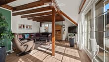 Bright living room with wooden beams, large windows, a dark brown sectional sofa, a coffee table, a leather armchair, a TV, and a wooden cabinet.