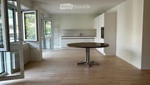 Open-plan space with light wooden flooring, white walls, and a single round table near the kitchen area. Large windows and sliding doors provide natural light and access to the outdoors.