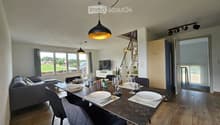 Open-plan living and dining area with a grey sofa, a dark dining table set for six, and a TV on a white stand. The space features wooden flooring, large windows with grey curtains, and modern pendant lights.