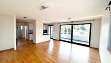 Bright, empty room with wooden flooring, white walls, and large sliding glass doors leading to a balcony. Minimalist design with recessed lighting and a small kitchen area visible in the background.