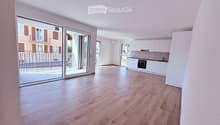 Bright, unfurnished living space with wooden flooring, white walls, recessed lighting, and a large sliding glass door leading to a balcony. Adjacent kitchen area with white cabinets and stainless steel appliances is visible.