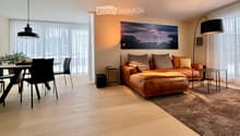 Modern living room with a brown leather sofa, coffee table, dining area with chairs, floor lamp, and large windows with sheer curtains.