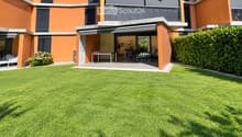 Modern house with orange and grey facade, large lawn, patio with outdoor furniture, and covered entrance area.