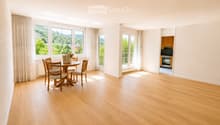 Bright, spacious room with wooden flooring, large windows with curtains, a dining table with chairs, and a handbag with flowers on it. A kitchen area is visible in the background.