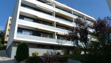 Modern multi-story apartment building with balconies, awnings, and surrounding greenery.
