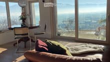 Bright living room with large windows offering a scenic view, featuring a brown leather sofa, throw pillows, a wooden coffee table, and a hanging pendant light.