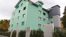 A modern residential building with three floors and several windows. The walls are painted green, and the roof has a triangular shape. The exterior is adorned with multiple white railings on the balcony, and there are bushes and trees on the sides. The bu