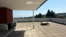 Roof terrace with tiled floor, low white railing, and a small square stone table. The area is covered by a large awning and offers a scenic view of the ocean and surrounding landscape.