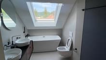 Modern bathroom with a large skylight, white bathtub, two sinks, toilet, and a round mirror.