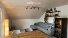 Modern living room with grey sectional sofa, wooden dining table, chairs, wine rack, shelves with bottles and plants, and a floor lamp.