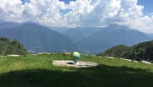 A grassy hilltop with a globe on a stone platform, overlooking a mountain range under a partly cloudy sky.