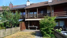 Two-story house with a tiled roof, solar panels, and a wooden balcony. The front yard includes a wooden fence, a small patio with a table and chairs, and lush greenery.