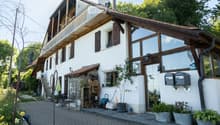 Two-story house with white walls, wooden accents, large windows, and a balcony on the upper level. The house features a stone chimney, a small garden area with potted plants, and a paved driveway.