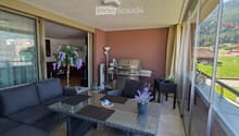 Balcony with a grey sofa, wicker table, chairs, flower pots, and a barbecue. Large windows offer a scenic view of the surrounding landscape.