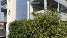 Modern two-story building with white and grey exterior, multiple balconies, and lush green bushes in front.