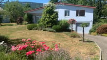 A single-story house with a red roof and light blue walls, surrounded by a well-maintained garden with various flowering plants, shrubs, and trees. The front yard features a gravel pathway leading to the house entrance.