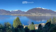 Panoramic view of a lake surrounded by snow-capped mountains, with trees and a small settlement visible on the shore.