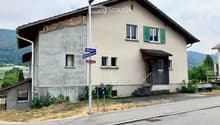 Two-story house with beige walls, green shutters, and a wooden entrance. The roof shows exposed wooden beams on one side. A street sign reading "Rehgasse" is visible in front.