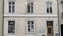 Two-story building with beige textured facade, multiple windows, decorative window frames, and a wooden front door. A white car and a scooter are parked in front.