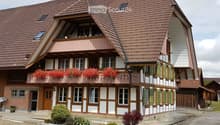 Two-story traditional house with red-tiled roof, white and brown facade, balcony with red flowers, green shutters, and small garden area.