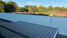 Modern balcony with a wooden table, glass railing, and a view of trees and buildings.