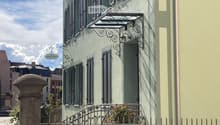 Building. A two-story building with light green walls, multiple windows with shutters, a decorative wrought iron balcony, and a small garden area behind a metal fence.