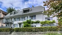 A multi-story residential house with a grey roof, multiple windows, and a balcony on the upper floor. The house is surrounded by greenery and has a stone pathway leading to the entrance.