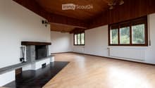 Spacious, unfurnished room with wooden floors, white walls, large windows, and a stone fireplace. The room features exposed wooden beams on the ceiling and radiators along the walls.