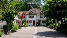 Two-story house with red shutters, white walls, and a tiled roof. The house features a driveway, garden, and is surrounded by trees and greenery.